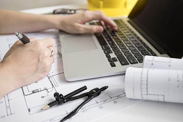 Digitaler Bauantrag - Stadt Forchheim Baupläne und ein Laptop befinden sich auf einem Tisch. Eine Person zeichnet an einem Plan und tippt zeitgleich am Laptop. Im Vordergrund liegt ein Zirkel.