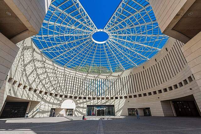 Eine modern eingfasste Piazza mit einer kunstvollen Metallüberdachung durch die ein blauer Himmel sichtbar ist.