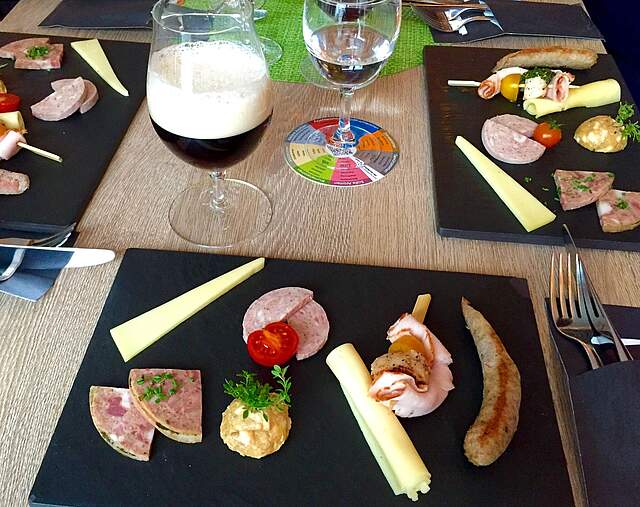 Drei Platten mit schön angerichtete Brotzeit sowie ein Glas Bier und ein Glas Wein auf einem Tisch.