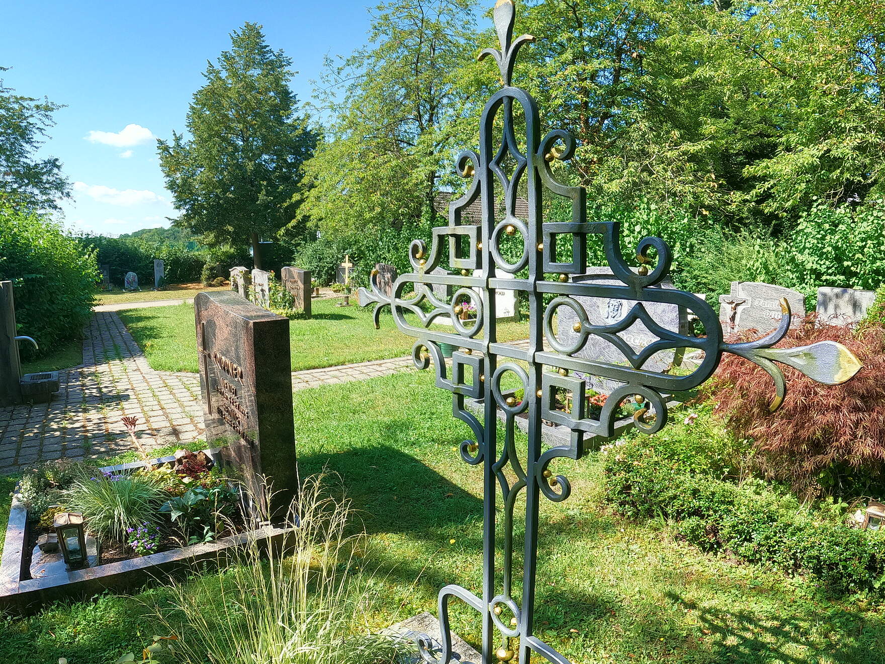 Bunt bepflanzte Erdgräber auf einem Friedhof. Im Vordergrund befindet sich ein Grabmal aus geschwungenem Metall. Das Areal ist umgeben von großen Bäumen.