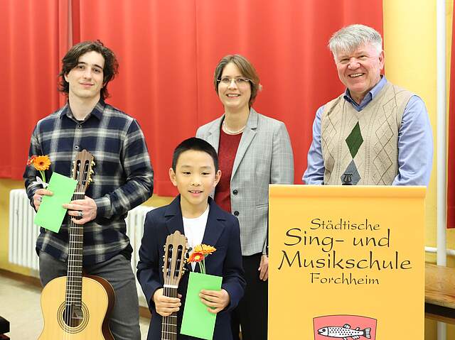 Die beiden Gewinner des Wettbewerbs mit Bürgermeisterin Dr. Annette Prechtel und dem Leiter der der Städtischen Sing- und Musikschule, Jürgen Hiltl vor einem roten Theatervorhang. Rechts im Bild steht ein auf einem gelben Plakat Städtische Sing- und Musikschule und ein Teil des städtischen roten Wappens mit Forellen.