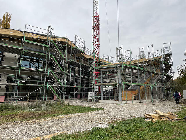 Der Rohbau der neuen Kita Merowingerstraße ist umgeben von Baustellengerüsten. In der Mitte steht ein Baukran. Im Vordergrund wechseln sich Wiesen- und Schotterflächen ab. Rechts vorne im Bild liegt ein Stapel mit Holzbaumaterial.