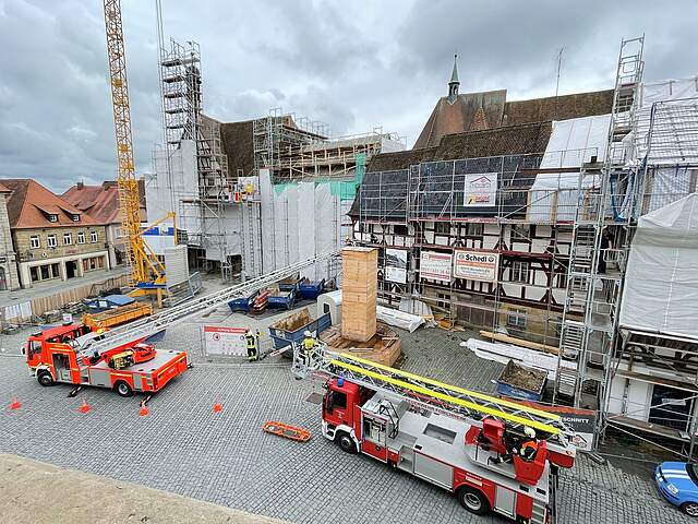 Zwei Feuerwehrautos mit Drehleiter parken vor dem eingerüsteten Fachwerkensemble und dem historischen Rathaus auf dem kopfsteingepflasterten Platz. Eine Drehleiter ist ausgefahren und hebt einen Feuerwehrmann auf Höhe der Holzeinhausung des Rathausbrunnens. Der Himmel ist grau und bewölkt. Auf dem Rathausplatz befindet sich noch ein Baustellenkran in der linken Bildhälfte.