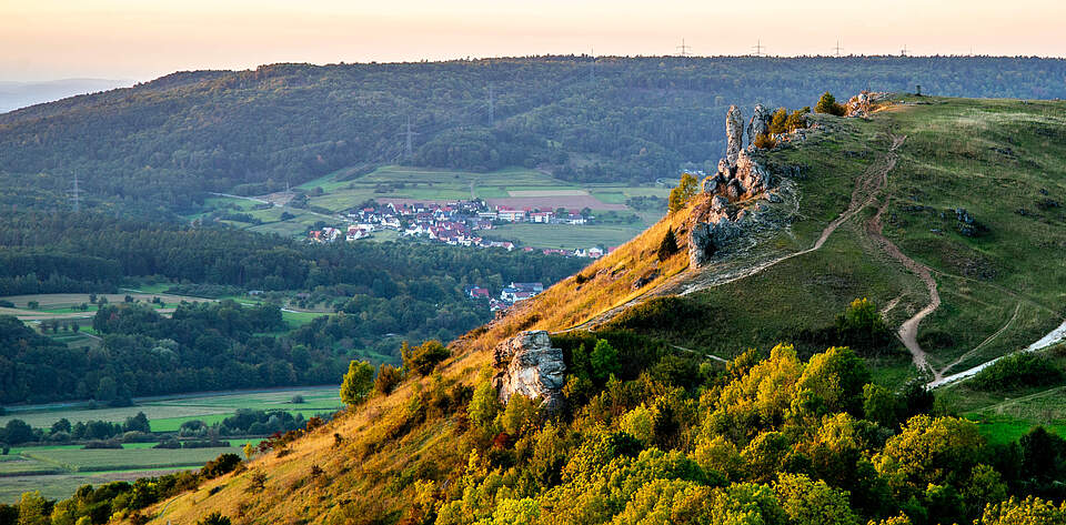 Schrägbildaufnahme von einem Berg bei Sonnenuntergang. Im Hintergrund befindet sich befindet sich ein Dorf, eingerahmt von Wald.