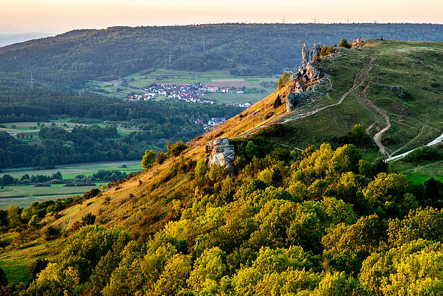 Schrägbildaufnahme von einem Berg bei Sonnenuntergang. Im Hintergrund befindet sich befindet sich ein Dorf, eingerahmt von Wald.