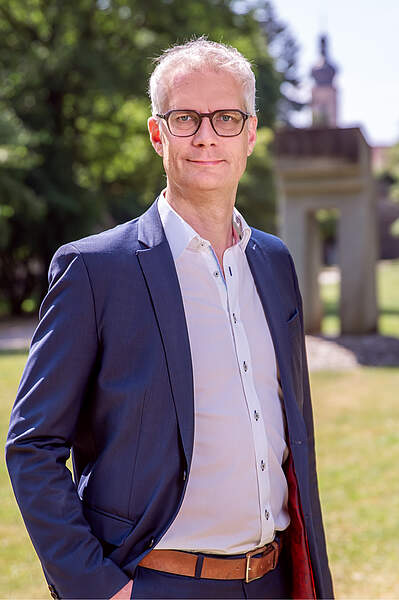 Oberbürgermeister Dr. Uwe Kirschstein steht bei Sonnenschein im Stadtpark. Die rechte Hand hat er locker in die Hosentasche gesteckt. Im Hintergrund sieht man die Kirchturmspitze der Martinskirche und ein Denkmal.