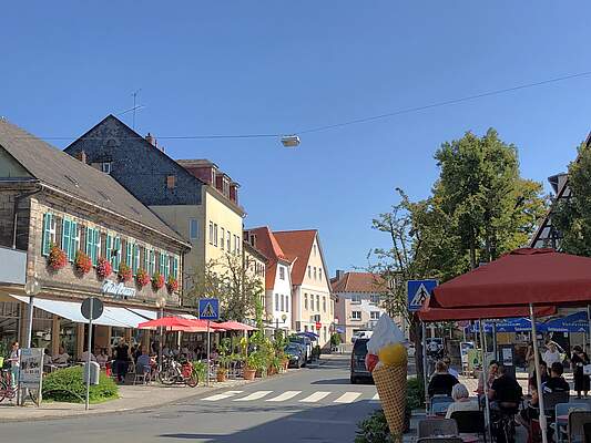 Straßenzug in der Forchheimer Innenstadt im Sommer. In der Mitte verläuft eine Straße mit einem Zebrastreifen. Auf beiden Seiten befinden sich gut besuchte Gastronomien und sommerliche Bepflanzung mit Blumen.