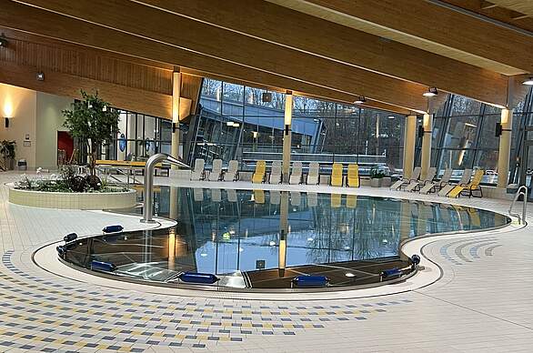 Ein Entspannungsbecken in einem Freizeitbad mit Wasserfalldüse am Beckenrand und einer Liegestuhlreihe entlang der Glasfront im Hintergrund.