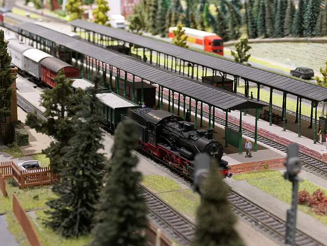 ine Modelleisenbahn-Landschaft zeigt einen Zug mit einer Dampflok an einem Bahnhof stehen. Im Hintergrund befinden sich auf einer Straße Pkws und zwei Lastwagen.