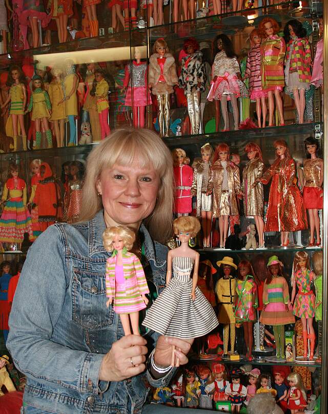 Eine Frau hält zwei Barbie-Puppen in den Händen. Hinter ihr befinden sich mehrere Glasvitrinen mit vielen unterschiedlichen Barbiepuppen.