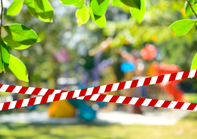 Im Hintergrund sieht man schemenhaft einen Spielplatz. Im Vordergrund ist gekreuzt ein Absperrband angelegt und darüber der Text "Spielplatz kurzzeitig geschlossen.