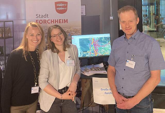 Corinna Stirnweiß, Leiterin des Stadtplanungsamtes (links), Geomatikerin Lisa Kaletsch und Projektleiter Matthias Hoffmann vom Stadtplanungsamt am Info-Stand der Stadt Forchheim. Im Hintergrund steht ein Roll-up mit einer Luftaufnahme der Stadt Forchheim und dem Stadtwappen. Daneben sieht man einen Bildschirm mit der Kartenansicht der Stadt und den Messungspunkten der Floating Car Data Analyse.