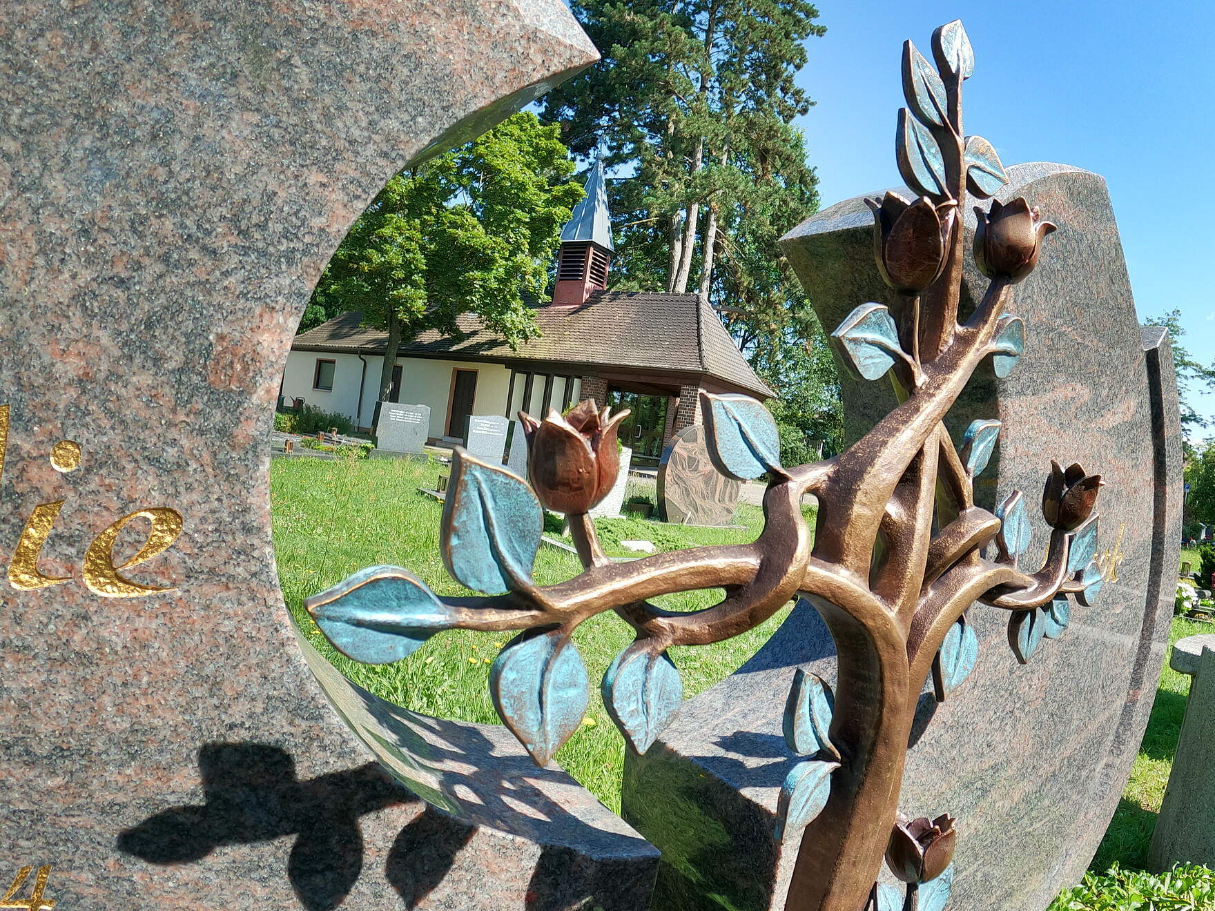 Blick durch einen Grabstein mit einer Rosenskulptur auf die eine kleine Leichenhalle an einem Friedhof.