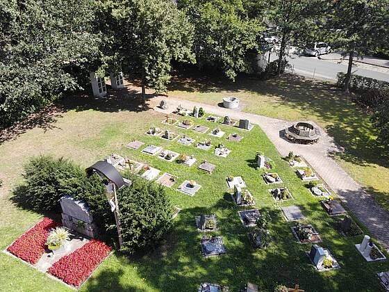 Luftbildaufnahme von einem Friedhof mit bunt bepflanzten Gräbern, einem Dankmal für Kriegsgefallene und einer runden Sitzgruppe. Das Areal ist umgeben von großen Bäumen.