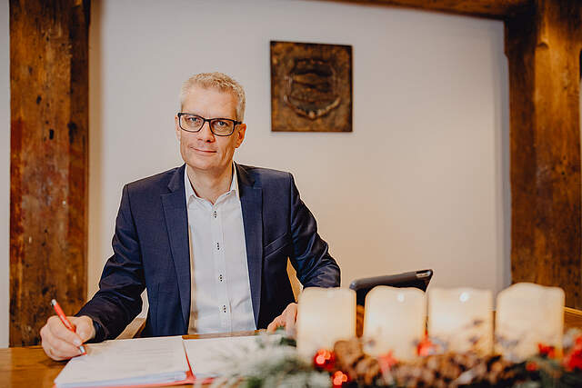 Ein Mann im Anzug sitzt an einem Schreibtisch und blickt in die Kamera. Auf dem Tisch liegen Dokumente. Ein Adventsgesteck mit vier brennenden Kerzen befindet sich ebenfalls auf dem Schreibtisch im Vorgrund. An der weißen Wand im Hintergrund sieht man das Forchheimer Stadtwappen mit den beiden Forellen.