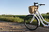 Ein Kiesweg führt durch eine Wiesenlandschaft. Man sieht einen blauen Himmel und ein weißes abgestelltes Damenfahrrad mit einem Fahrradkorb am Lenker.