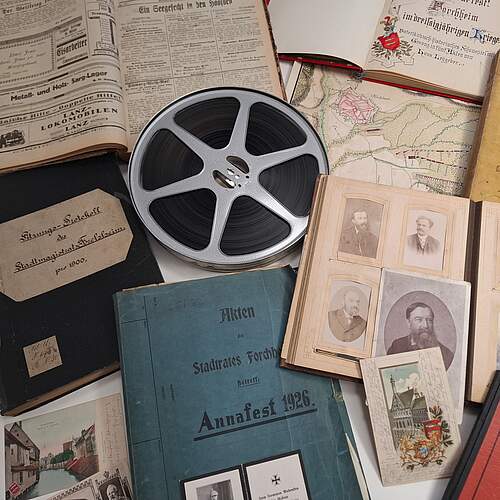 Verschiedene Archivalien wie Tonband, aufgeklappte Fotoalben und Akten liegen als Collage angeordnet auf einem Tisch.