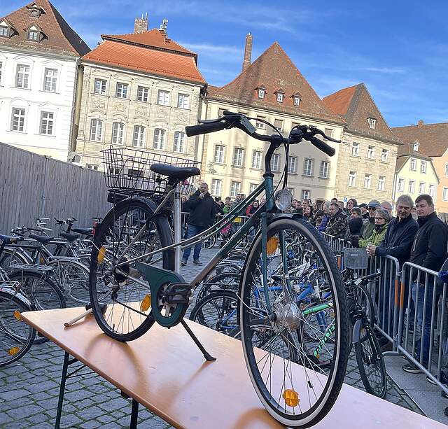 Ein Fahrrad wird auf einem Tisch bei einer Versteigerung auf einem Stadtplatz präsentiert. Im Hintergrund sieht man interessierte Personen an Absperrgeländern und mehrere Fahrräder sowie eine Häuserzeile bei sonnigem Wetter.