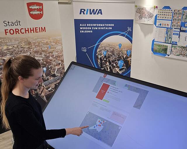 Eine Frau zeigt an einem Digitalen Tisch (Monitor) auf einen Lageplan.