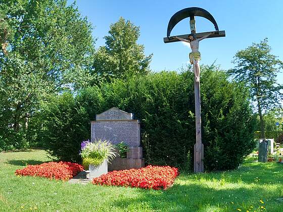 Ein Gedenkstein an die Weltkriegopfer unterflanzt mit roten Sommerblumen und einer Blumenschale in der Mitte vor einem großen Busch. Links steht ein großes Kreuz. Der Gedenkstein ist umgeben von einer Rasenfläche. Im Hintergrund befinden sich Bäume.