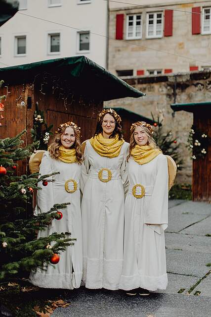 Die Forchheimer Weihnachtsengel 2025 Drei als Engel verkleidete junge Frauen stehen vor festlich geschmückten Weihnachtsbuden.