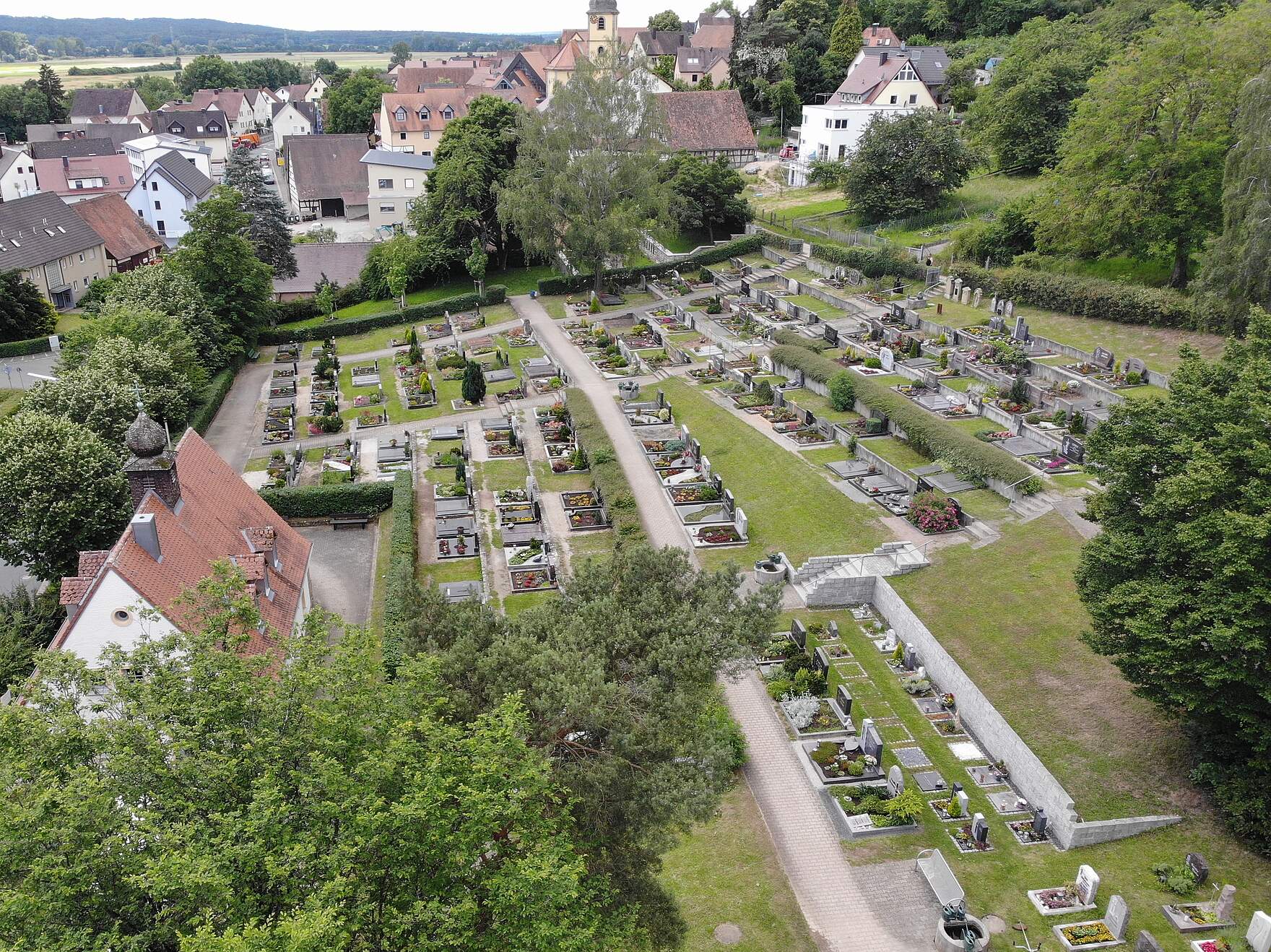 Ein terrassenförmig angelgeter Friedhof mit bunt bepflanzten Erdgräbern. Eine Treppe verbindet die einzelnen Ebenen. Umrahmt wird das Areal von hohen Bäumen. Links erkennt man hinter einer Baumkrone das Dach der Aussegnungshalle. Im Hintergrund befinden sich Wohnhäuser.