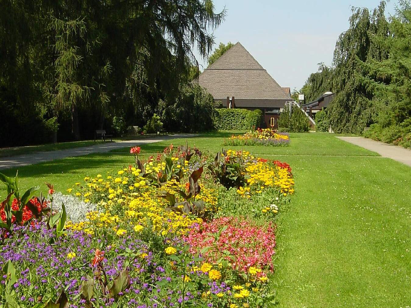 Aussegnungshalle im Neuen Friedhof (Heimgartenstraße) Aussegnungshalle im Neuen Friedhof aus den 80er Jahren. Im Vordergrund befindet sich eine große Rasenfläche mit einer sommerlich bepflanzten Staudenrabatte.