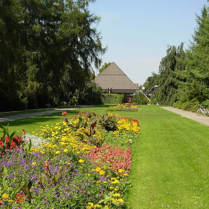 Aussegnungshalle im Neuen Friedhof aus den 80er Jahren. Im Vordergrund befindet sich eine große Rasenfläche mit einer sommerlich bepflanzten Staudenrabatte.