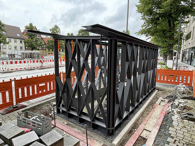Hochbauten am Tiefagaragenabgang Süd am Paradeplatz Forchheim Ein moderner Hochbau aus dunklem Stahl wird gerade an einem Treppenabgang zu einer Tiefgarage errichtet. Umliegend befindet sich eine Großbaustelle mit Absperrungen und verschiedenem Baumaterial.
