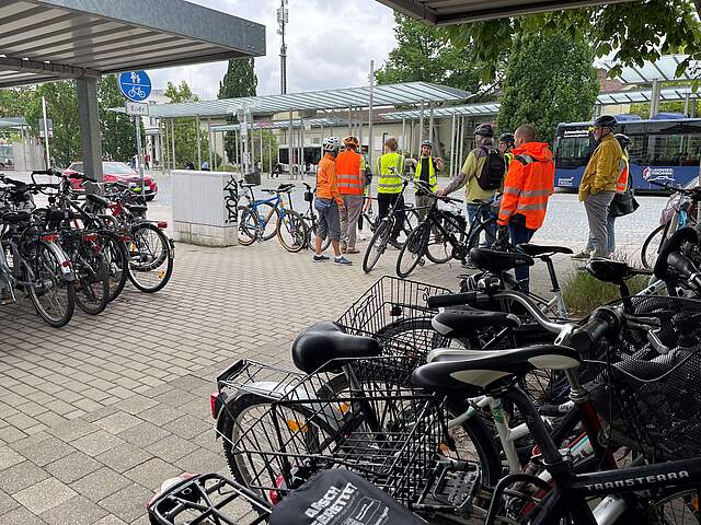 Radfahrergruppe mit Helmen sowie geleben und orangefarbenen Warnwesten stehen mit ihren Rädern an einem Fahrradabstellplatz vor einem Busbahnhof.