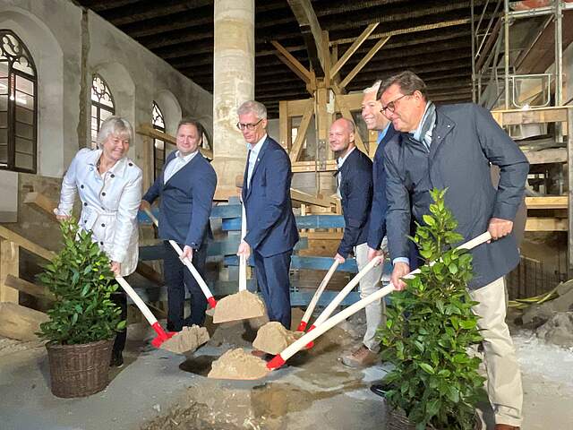 Eine Gruppe aus sechs Männern und Frauen halten jeweils eine mit Sand gefüllte Schaufel. Sie stehen in einem hohen, großen Raum, in dem sich eine Großbaustelle befindet.