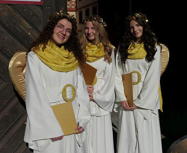 Drei junge Frauen in weiß-goldenen Engelskostümen mit goldenen Flügeln stehen nebeneinander vor einem großen Holztor.