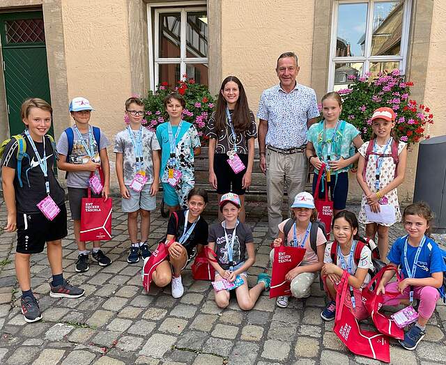 Kinder im Schulalter stehen in einer Gruppe mit Bürgermeister Udo Schönfelder vor dem Verwaltungsgebäude. Einige Kinder tragen Schirmmützen und rote Jutebeutel mit der Aufschrift "Entdecker Täschla".