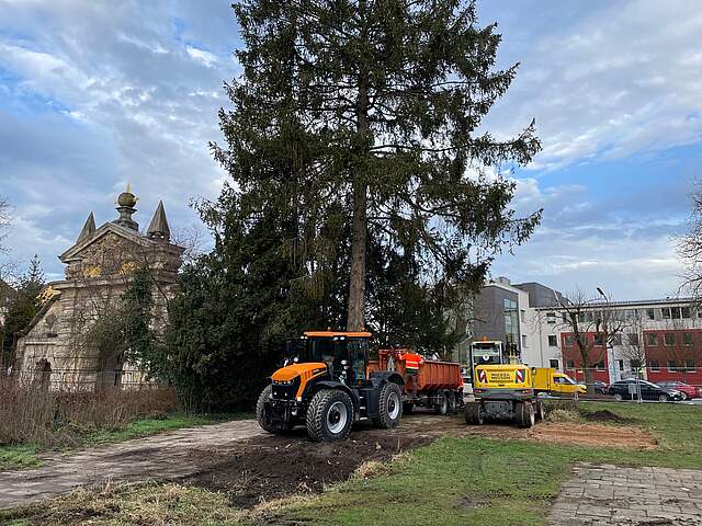 Ein Traktor mit Anhänger parkt neben einem Bagger auf einer Grünfläche ,it großen Bäumen in einer Stadt. Links im Bild befindet sich ein historisches Stadttor. Im Hintergrund sieht man ein Bürogebäude.