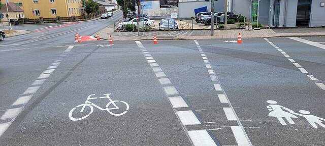 Weiße Straßenmarkierungen zeigen einen Fußgängerüberweg und einen in eine Straße einmündenden Fahrradweg mit einer roten Warnmarkierung an der Einmündung.