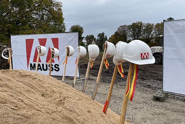 Ein großer Sandhaufen mit acht Spaten, die mit Bändern in den Forchheimer Farben gelb und rot geschmückt sind. Auf den Spaten stecken weiße Baustellenhelme. Im Hintergrund befinden sich zwei weiße Bauzäune mit dem rot-weißen Logo der Firma MAUSS BAU.