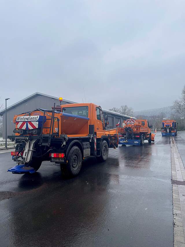 Drei große Räum- und Streufahrzeugs fahren auf einer Straße bei trüben Wetter hintereinander.