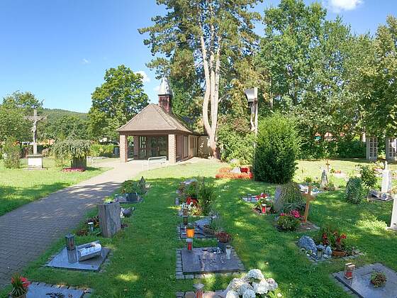 Urnengräber mit Blumeschmuck umgeben von Rasenflächen auf einem Friedhof. Links im Bild führt ein gepflasterter Weg zum Leichenhaus mit Backsteinsäulen und einem Glockenturm. Große Bäume umrahmen das Areal.inen 