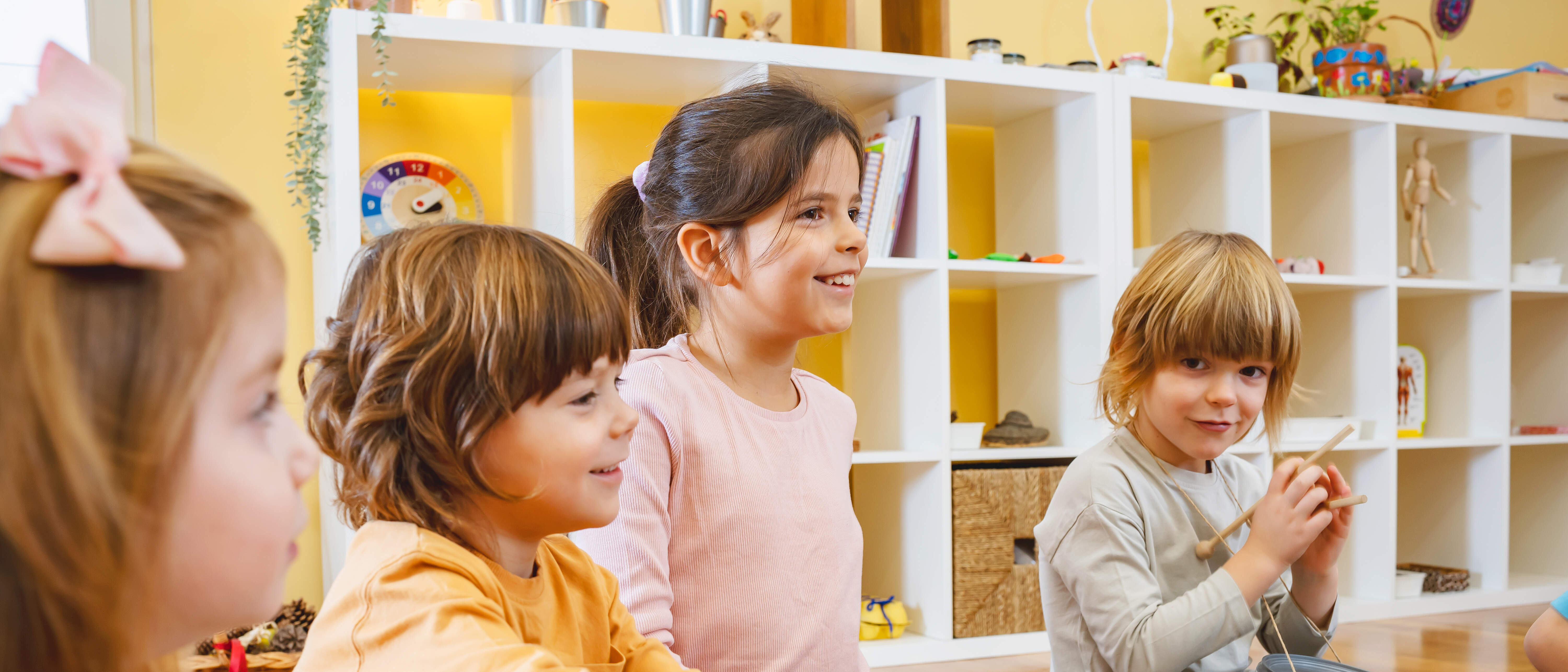 Kindergarten Morgenkreis Vier Kindergartenkinder mit verschiednenen Musikinstrumenten sitzen nebeneinander auf dem Boden eines Kindergartenraumes. Im Hintergrund befindet sich ein weißes Regal mit Spielsachen.
