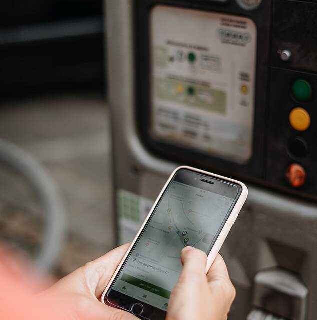 Eine Person steht mit einem Mobiltelefon in den Händen vor einem Parkautomaten.