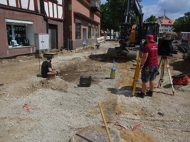 Nach dem Abbruch einer Belagsfläche an einem großen Stadtplatz untersuchen zwei Arbeiter den Boden nach archäologischen Funden.