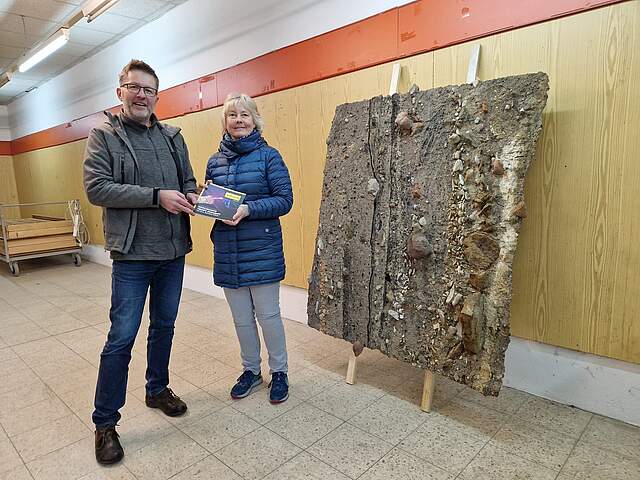 Ein Mann und eine Frau stehen links neben einem sogenannten Sedimenttransferpräparat, das an einer Wand lehnt. Das Präparat zeigt anhand von Gesteinsschichten anschaulich Forchheims Geschichte.