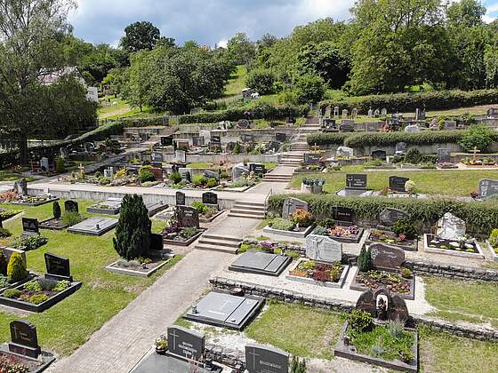 Ein terrassenförmig angelgeter Friedhof mit bunt bepflanzten Erdgräbern. Eine Treppe verbindet die einzelnen Ebenen. Im Hintergrund befinden sich hohe Bäume.