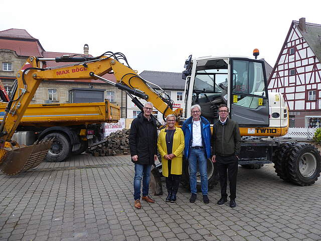 Drei Männer und eine Frau stehen vor einem Bagger und einem LKW an einer Baustelle.