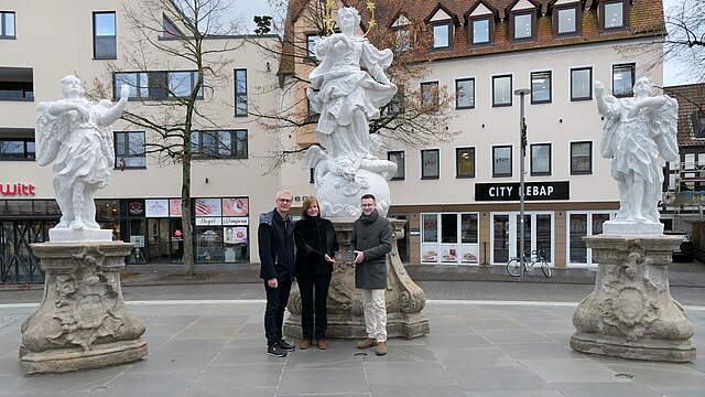 Drei Personen stehen bei einer historischen Figurengruppe auf einem Stadtplatz und präsentieren ein Messingschild mit der Aufschrift "Heimatverein Forchheim - Restauriert 2024 - gefördert vom Heimatverein Forchheim Ofr. e.V." .