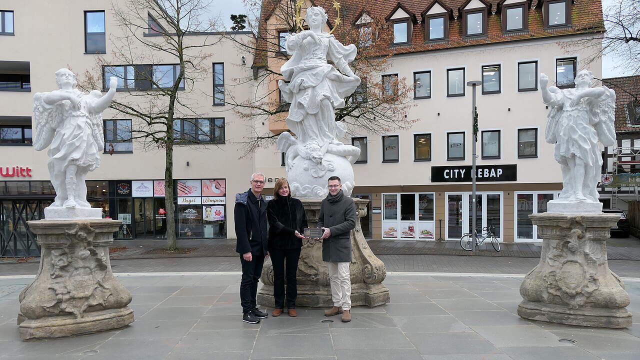 Drei Personen stehen bei einer historischen Figurengruppe auf einem Stadtplatz und präsentieren ein Messingschild mit der Aufschrift "Heimatverein Forchheim - Restauriert 2024 - gefördert vom Heimatverein Forchheim Ofr. e.V." .