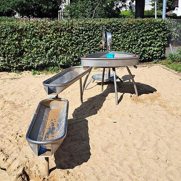 Wasserspielanlage in der Von-Ketteler-Straße Ein mehrteiliges Wasserspielgerät in einem Sandkasten auf einem Spielplatz.