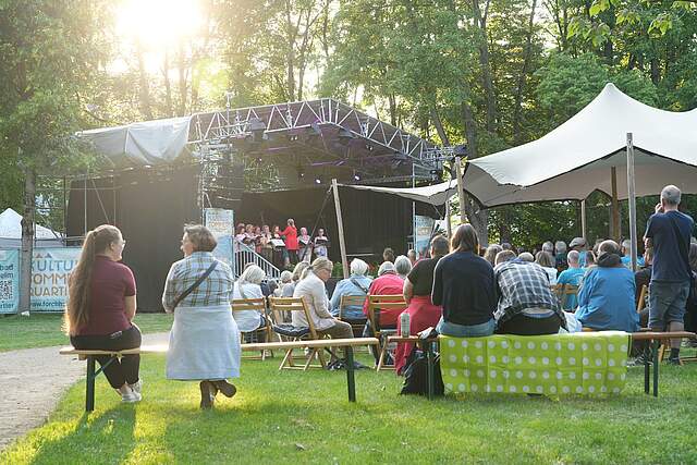 Zuschauer sitzen auf Bänken und Stühlen vor einer Bühne bei einem Open-Air Konzert im Grünen.