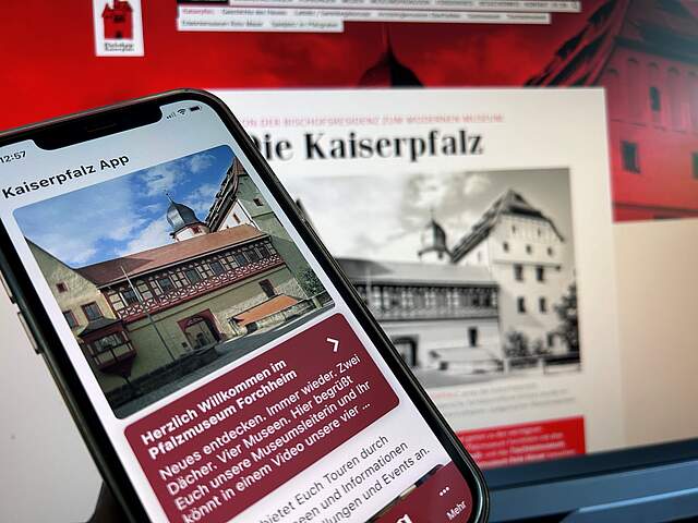 Im Vordergrund befindet sich ein Handy, das eine das eine App zeigt. Auf dem Display sieht man die Frontansicht einer Burg und unterhalb einen rot hinterlegten Text. Im Hintergrund sieht man verschwommen einen Computerbildschirm.