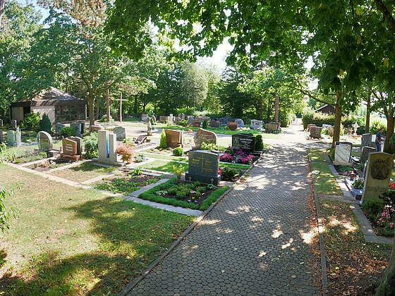 Bepflanzte Erdgräber auf einem Friedhof, teilweise unter schattigen Bäumen. Im Hintergrund befindet sich eine Leichenhallte und ein großes Kreuz. Die Anlage ist umgeben von großen Bäumen